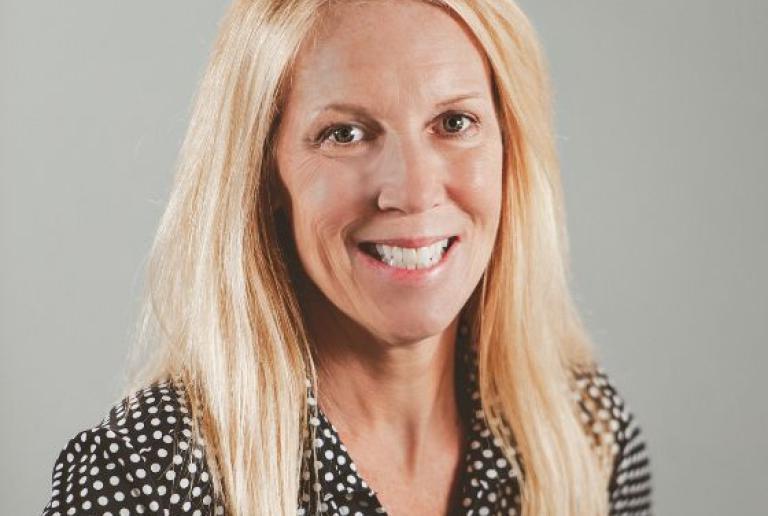 The image shows a woman with long, straight blonde hair wearing a black blouse with white polka dots. She has a friendly expression with a broad smile and is facing slightly to her right. The background is a plain light gray. She has light skin and an approachable demeanor.